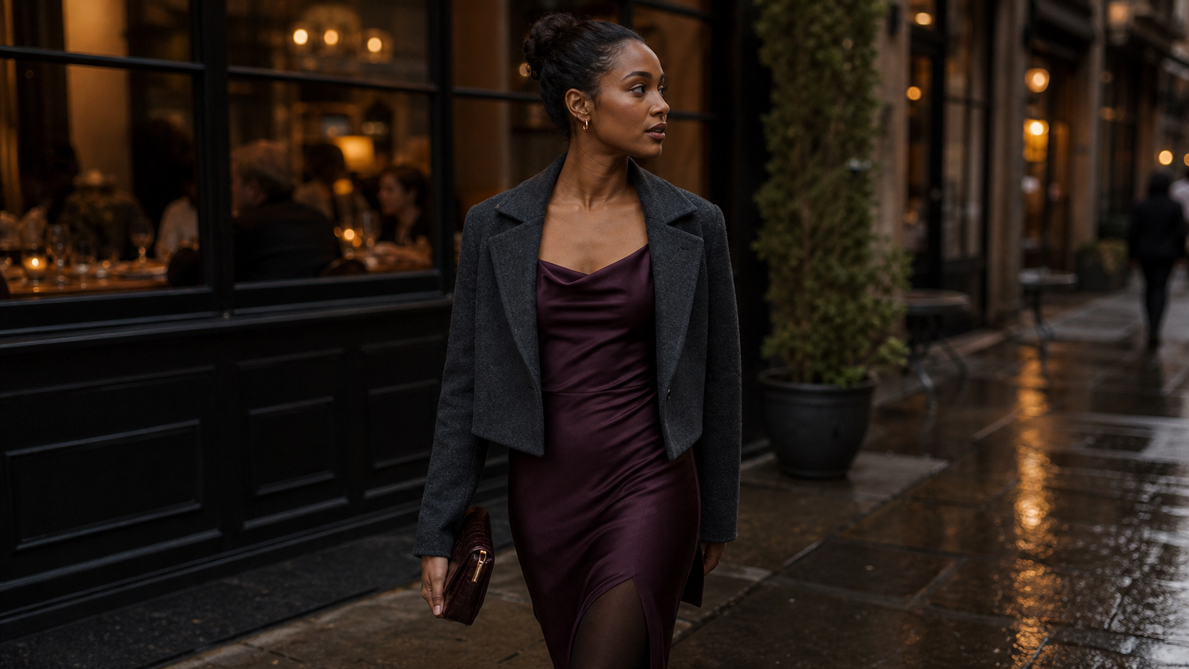 A guest in a plum satin dress and charcoal jacket outside a warm restaurant on a rainy fall evening.
