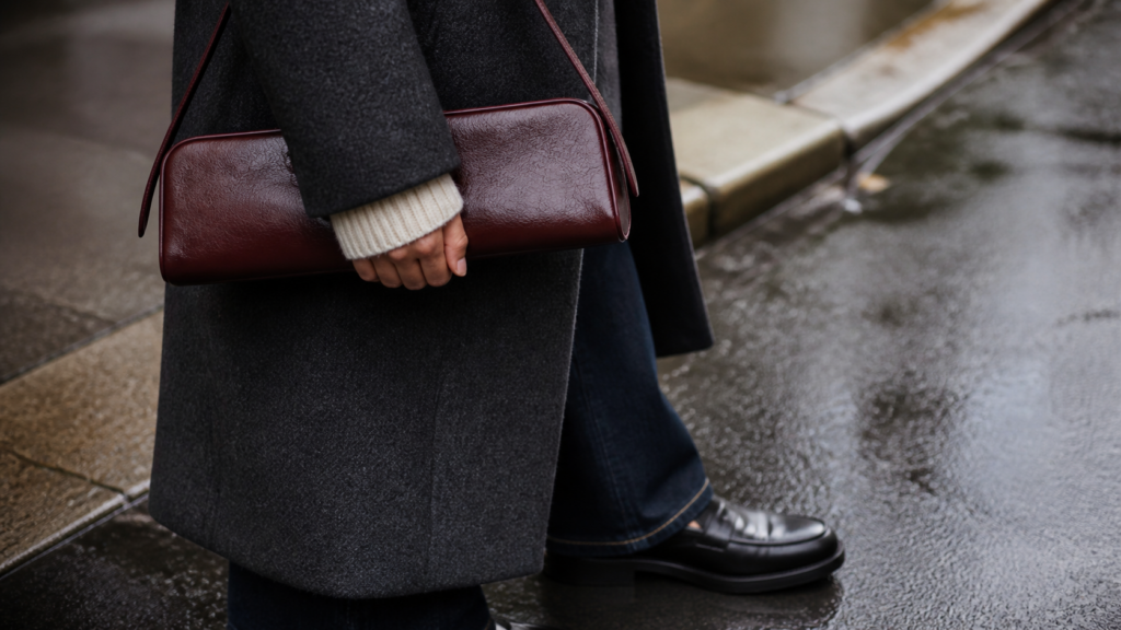 Oxblood east west shoulder bag styled with a charcoal coat and loafers on wet pavement.
